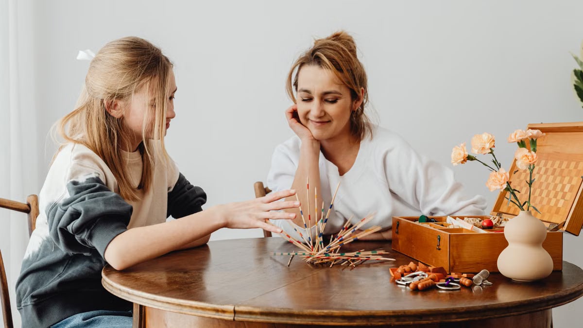 Two women enjoy a relaxed game at a wooden table, surrounded by craft supplies, flowers, and a decorative vase.